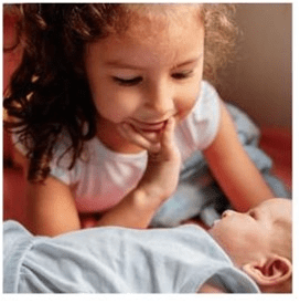 A young girl with curly hair is touched on the face by a baby wearing a blue jumper.