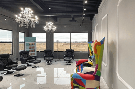 a photo of crave beauty. a colorful chair sits agains a white wall, and five leather recliners are sitting against windows opposite the chair