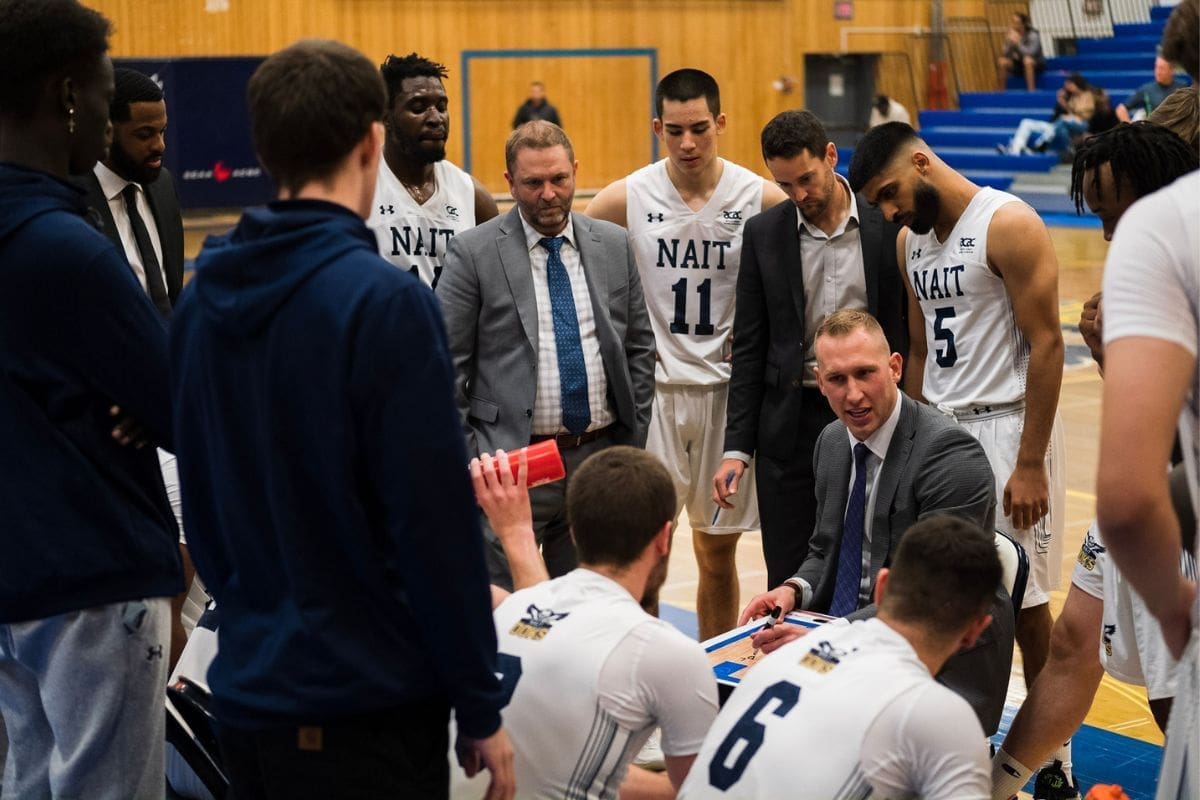 men’s bball team huddle The men's basketball team circles in a huddle. Coach Jordan Baker is in the middle, wearing a suit.