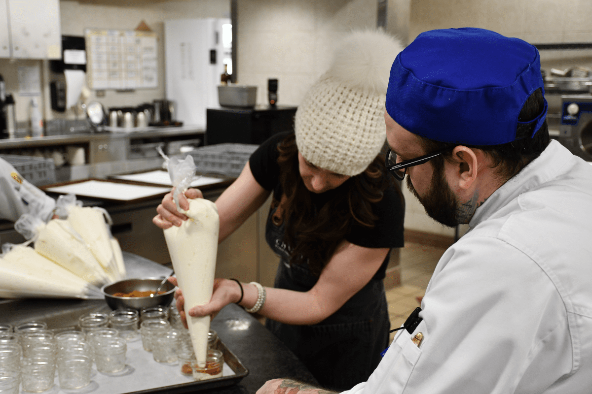 A woman holds a bag of whipped cream. A man stands behind her in a chef uniform.