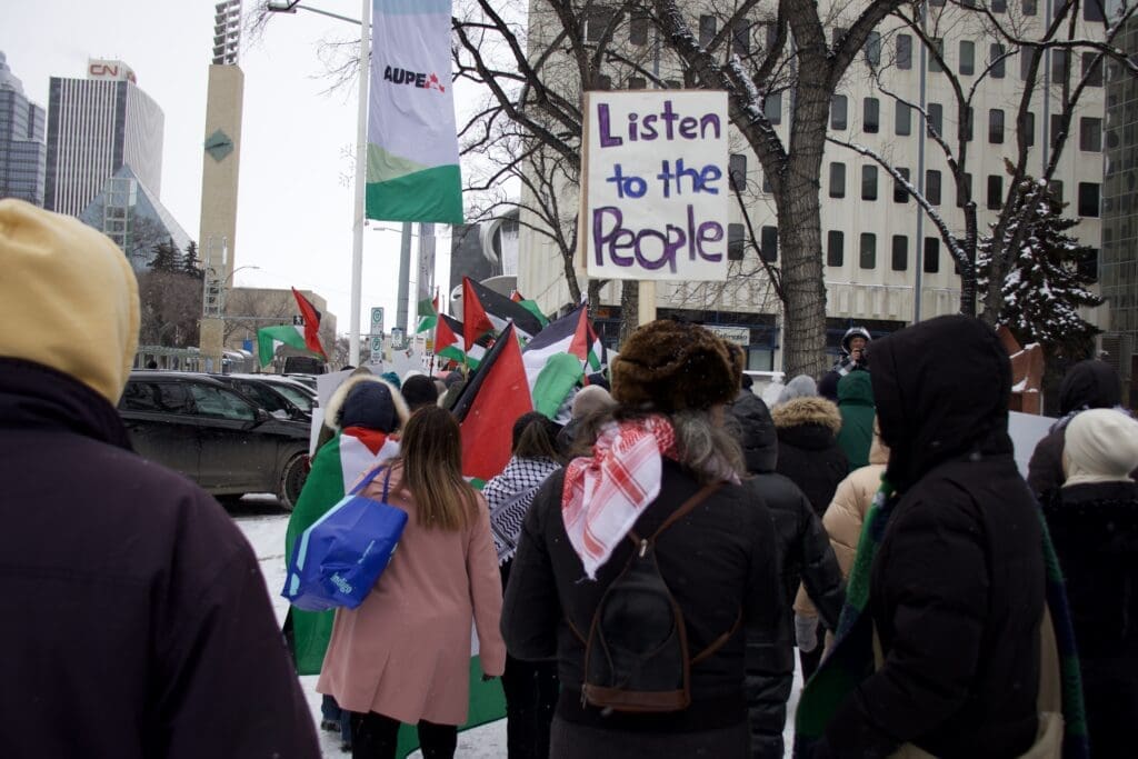 A crowd marches. Someone holds a sign that says "listen to the people." 