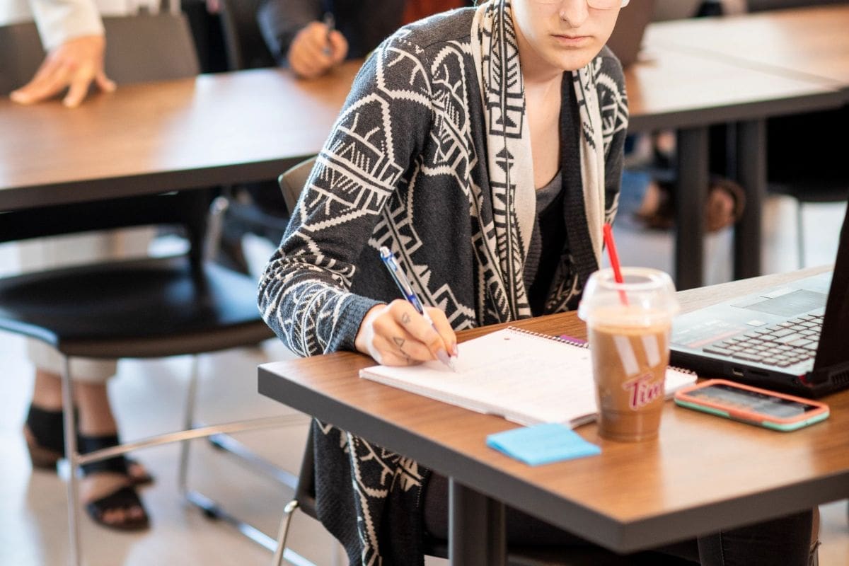bottom half of a person in a grey patterned sweater; they are writing in a notebook while sitting at a table in a classroom.