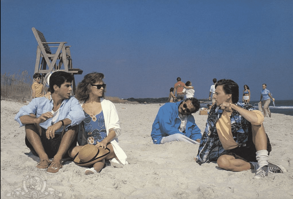 four people sit on a beach. one guy in the back looks like he is asleep. 