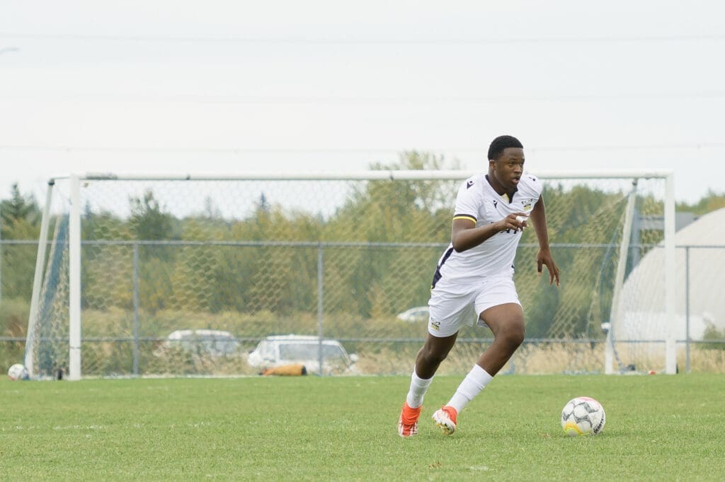 A dark-skinned man in a white soccer uniform runs down a soccer field with the ball at his feet.