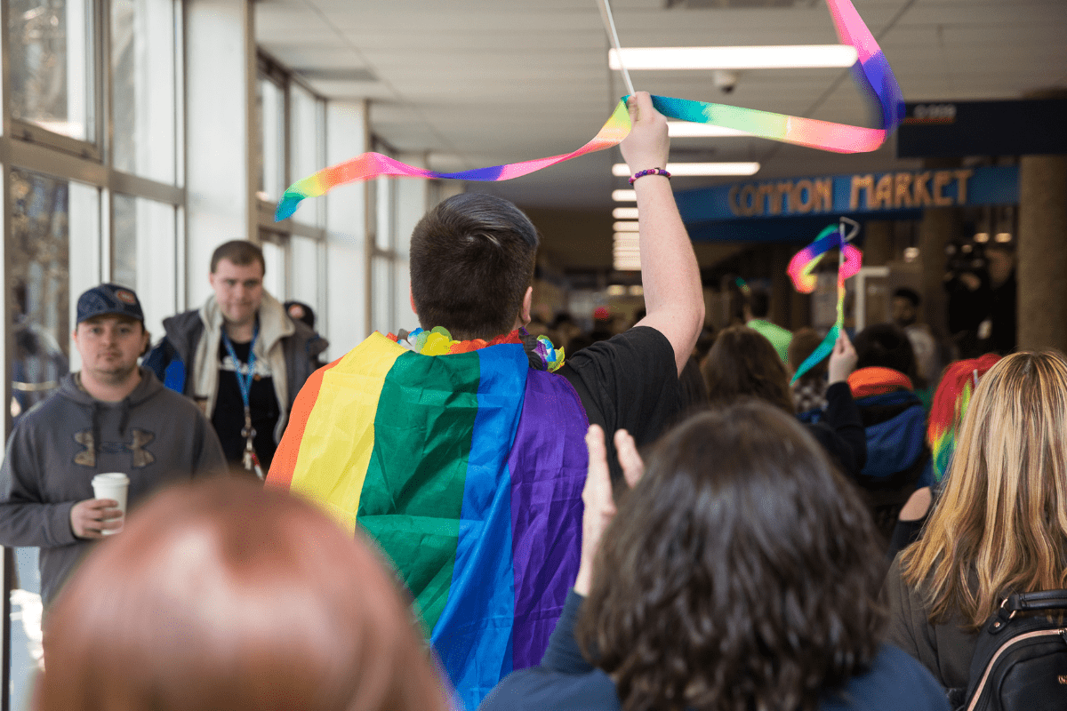 in the centre of a crowded hallway is a man with short brown hair wearing a rainbow flag and waving a rainbow streamer