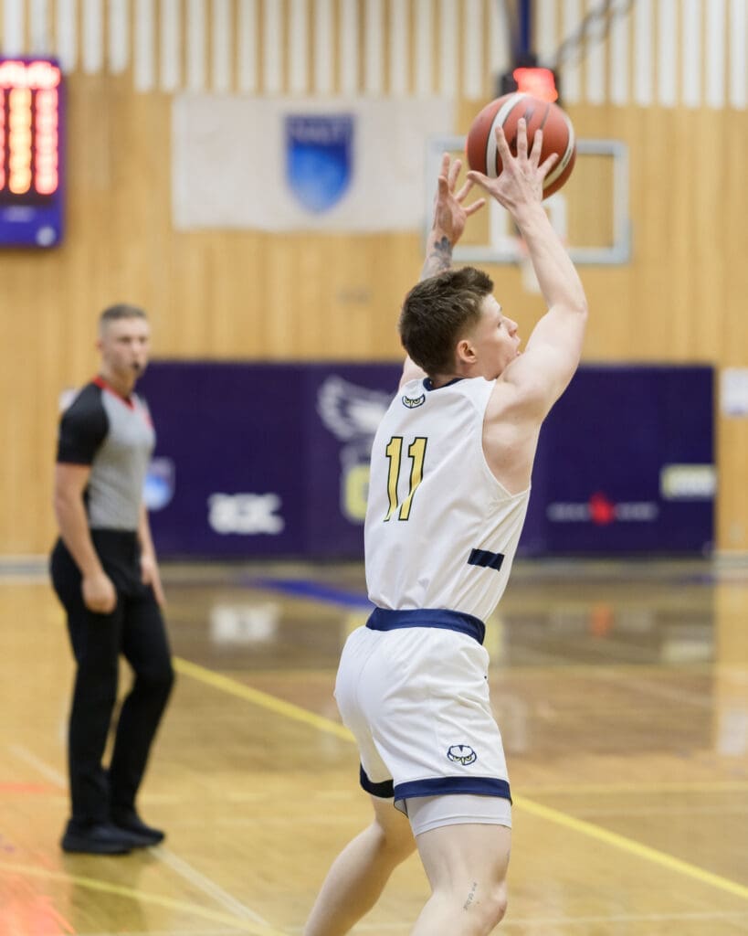 A white man wearing a white NAIT basketball uniform reaches up to catch a basketball. 