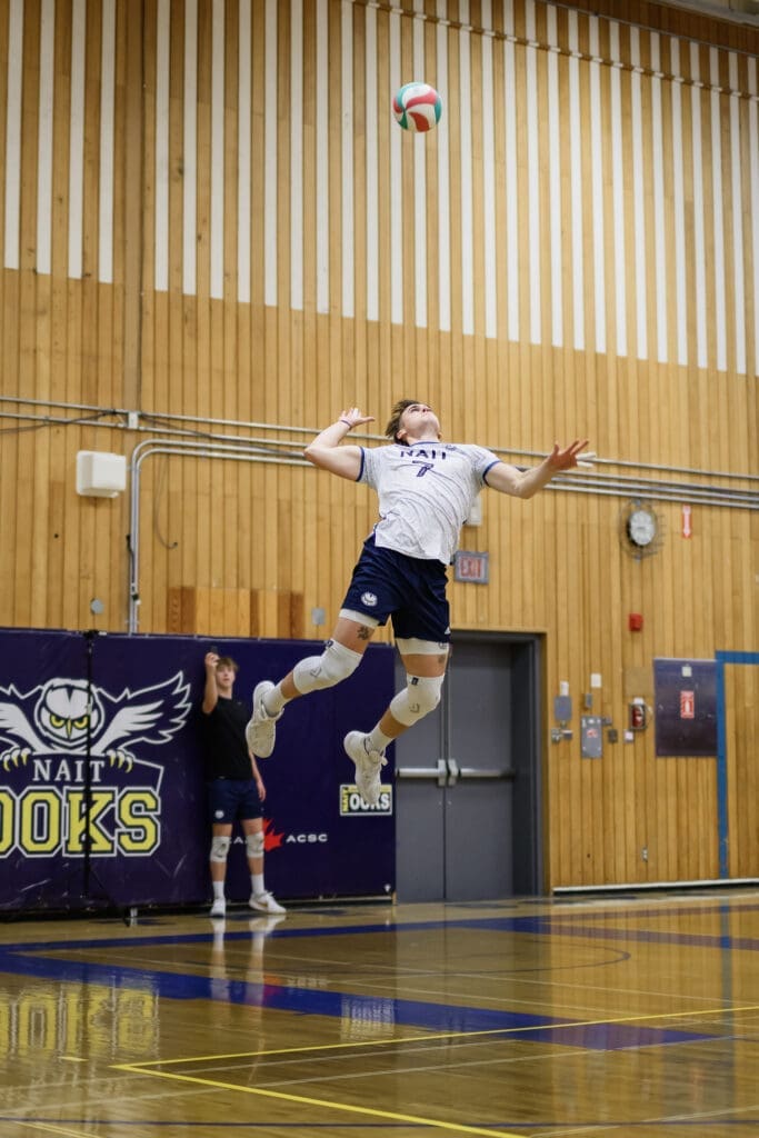 A white man in a blue and white volleyball uniform is jumping into the air to hit the volleyball. 