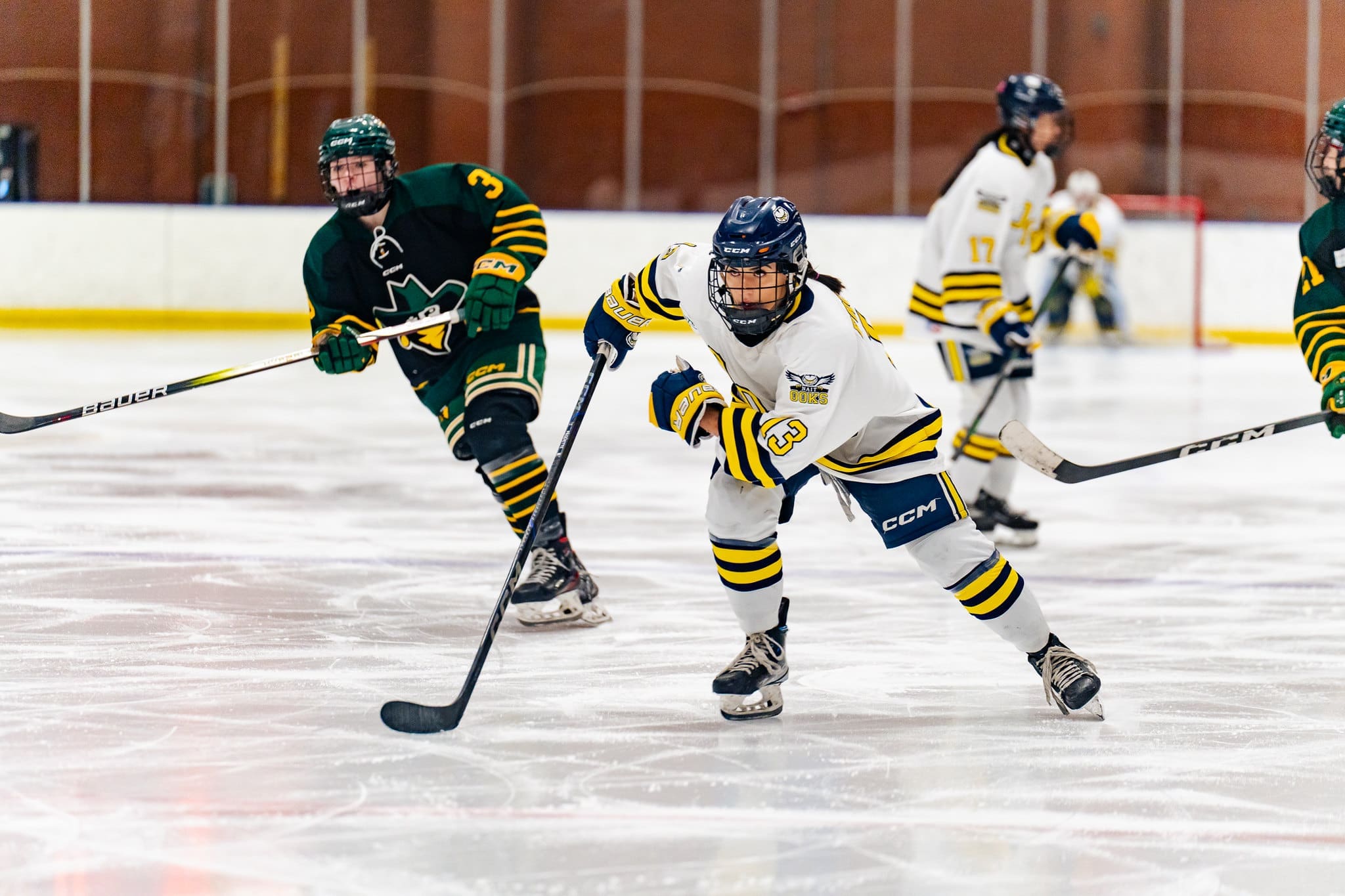 Female hockey players skate in a hockey rink. In the centre is Hanna Paquette, wearing a blue helmet and the white and yellow ooks uniform. 