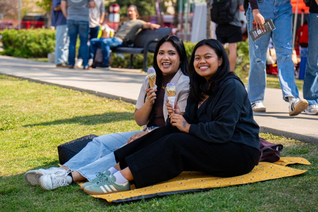 Two women sit on the grass and smile at the camera. They are holding ice cream cones. 