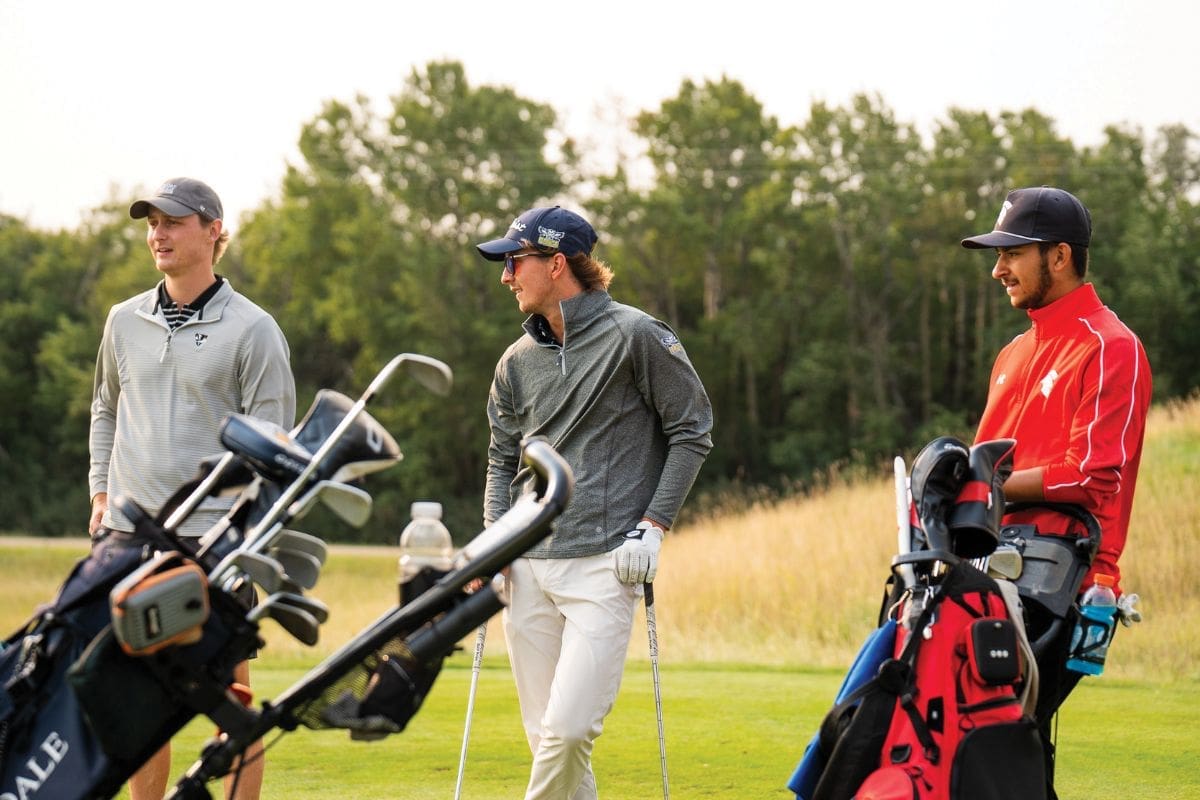 Three male golfers stand with their golf bags on a field.