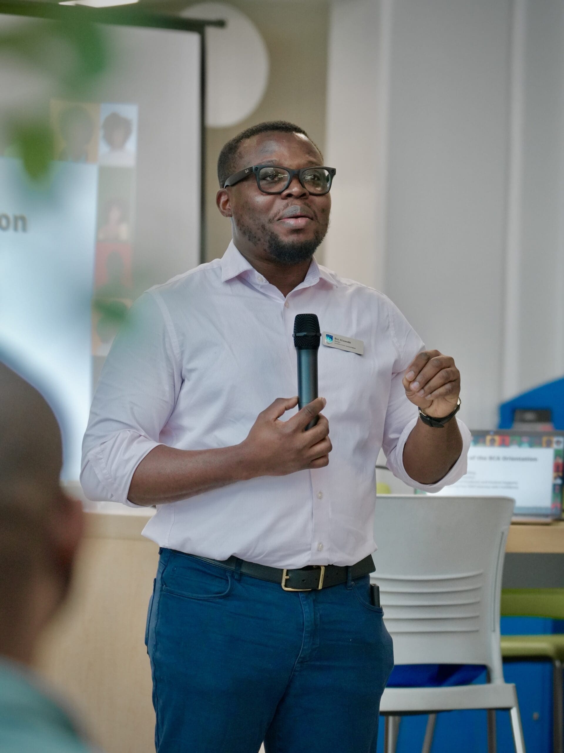 A Black man wearing glasses, a collared shirt and blue pants speaks into a microphone.