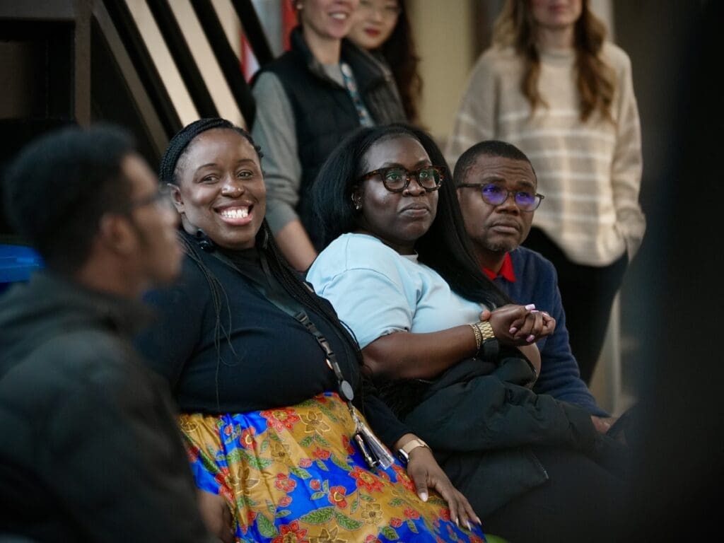 A Black woman smiles at something off-camera. She wears a black shirt and a colourful skirt. Two people sitting beside her stare thoughtfully.