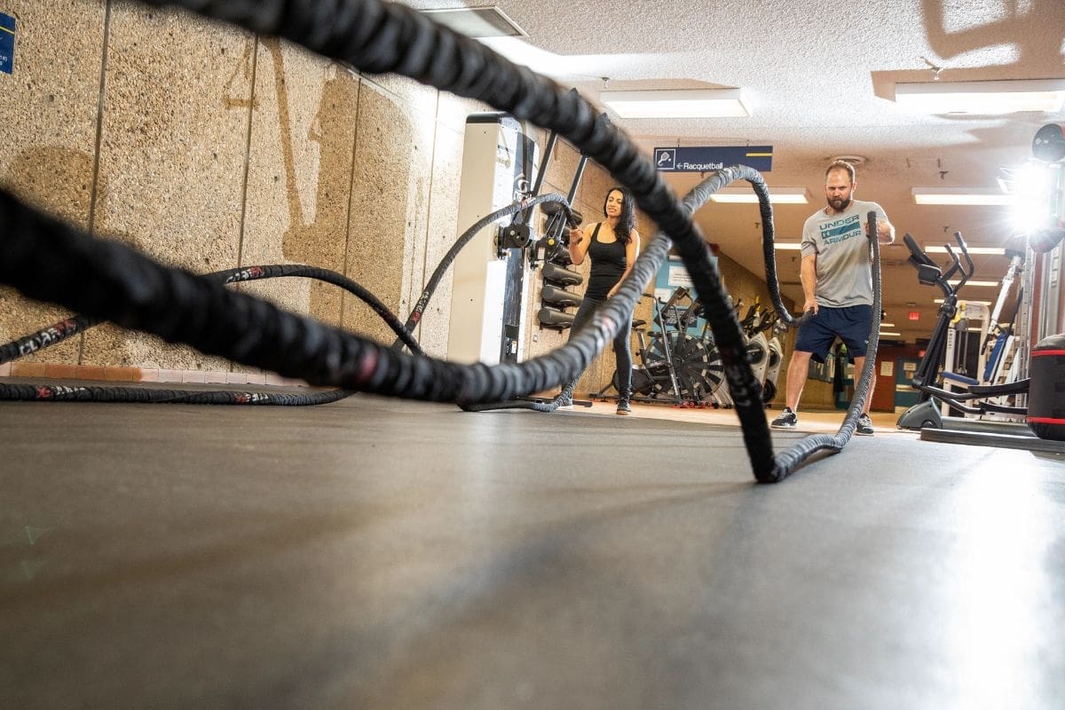 Thick fitness ropes swing in the air, with two people lifting them in the background.