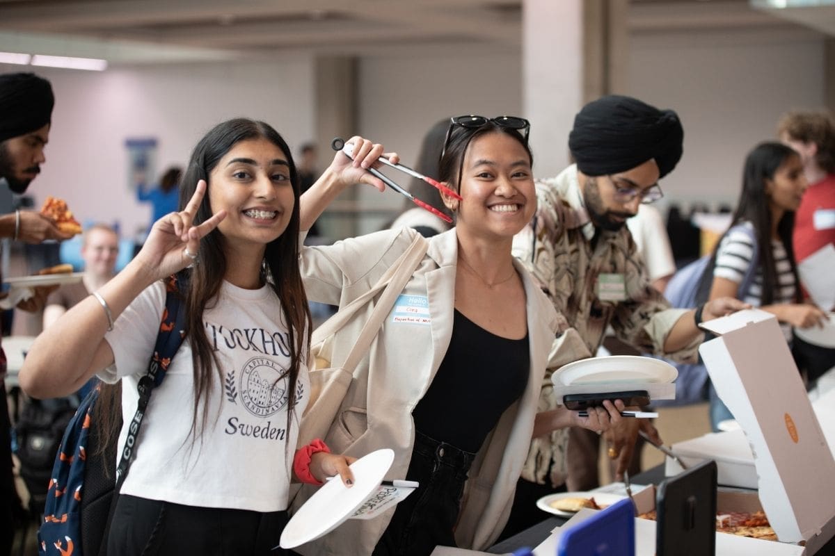 Two girls, one with long brown hair, and another with dark hair in a ponytail, smile and hold up peace signs to the camera. They are holding plates and waiting in a line.