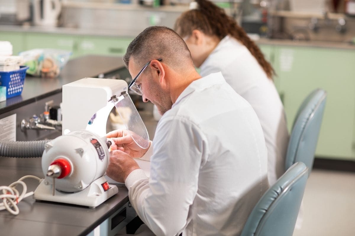 A man with short hair works on denture equipment. A person with a long brown ponytail sits behind him.