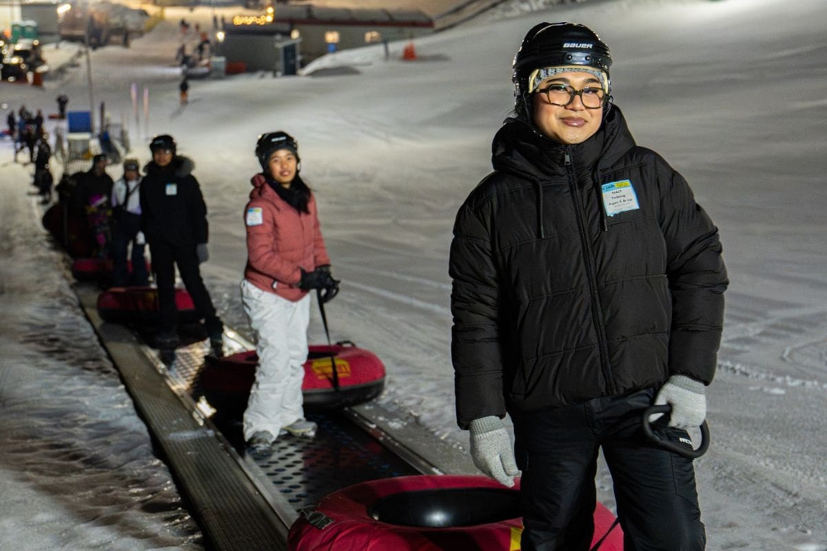 snow tubing A woman smiles at the camera. She is in winter gear and is at the front of a line. All are waiting to ride a tube down the ski hill.