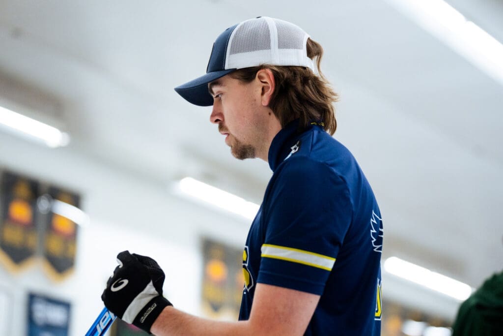 A man with shaggy brown hair looks to the left. He's wearing a white and blue hat and a blue short-sleeved sports polo. 
