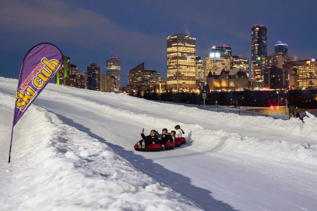 A group of smiling people in tubes slide down a ski hill. The city skyline is visible in the background.