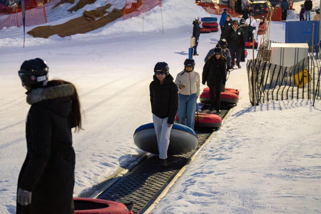 Young people in winter gear carry tubes up a snow hill.