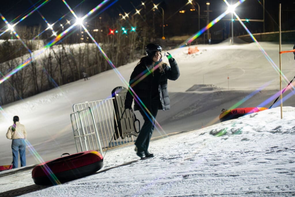 A girl in winter gear walks up a ski hill carrying a tube behind her. The light sparkles in the camera lens.