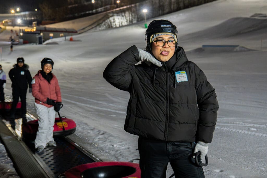A woman in a black winter coat and helmet sticks out her tongue as she carries a snow tube up a ski hill.