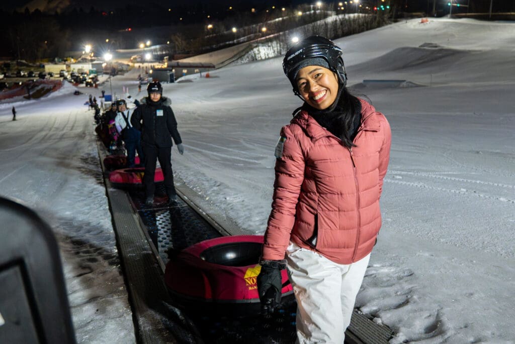 A girl in a pink coat and helmet smiles while holding a snow tube.