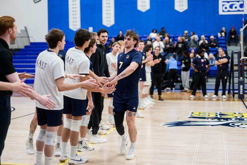 Male volleyball players form a line on the left. One male player shakes hands with a few of the players. 