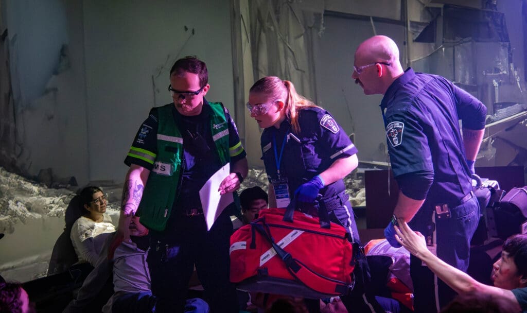 Three uniformed first responders examine a collapsed building scenario. One holds a large bag of supplies. In the background, a patient actor leans against rubble.