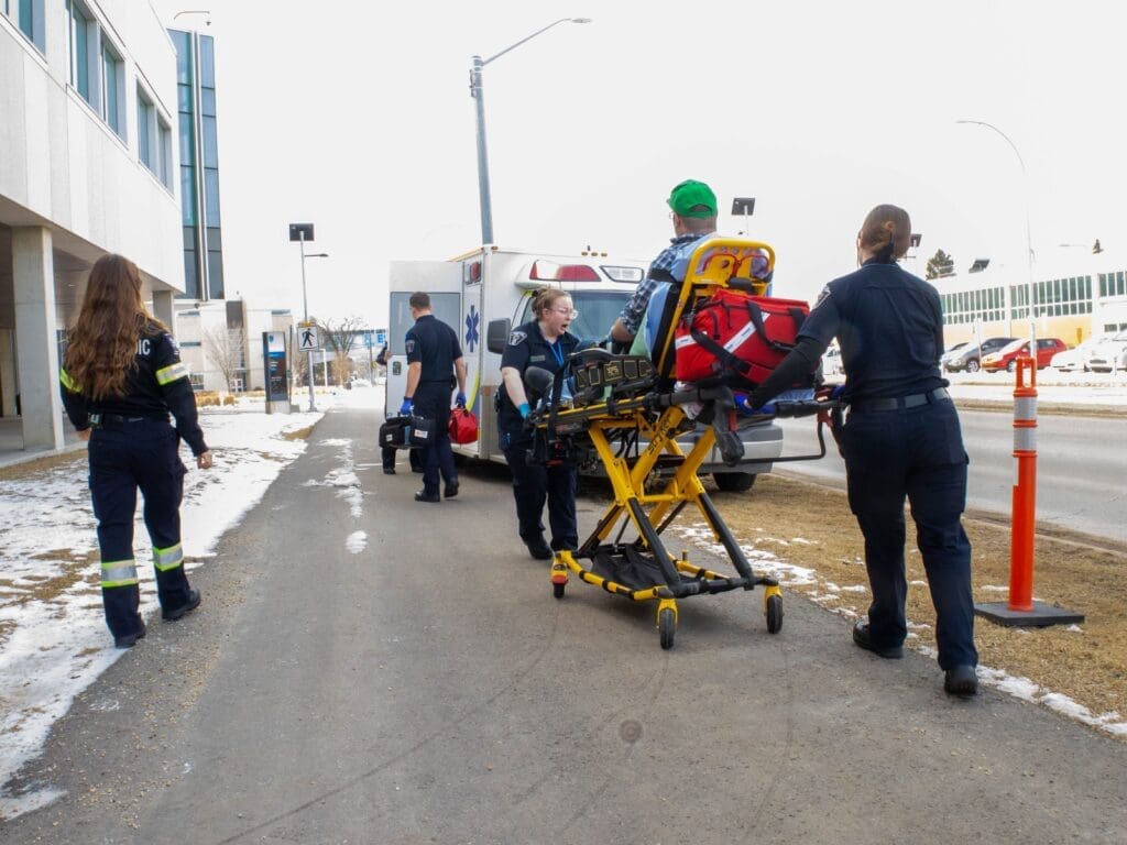 Four paramedic students transport a male patient on a stretcher outside NAIT campus. An ambulance is parked in the background.