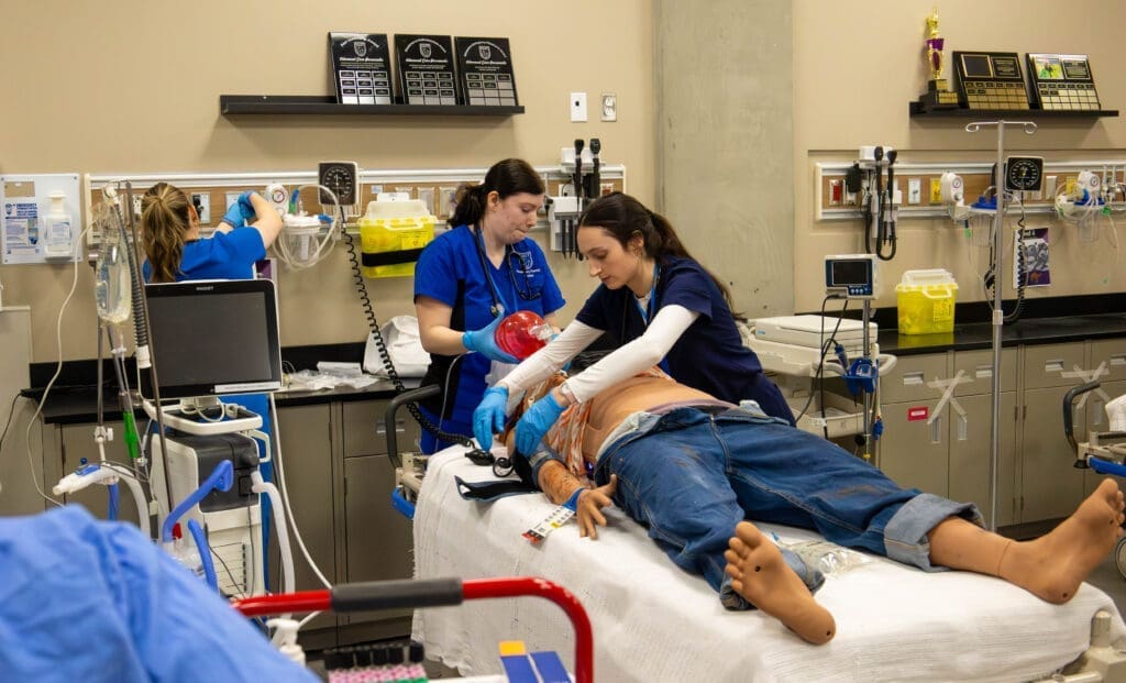 Two respiratory students, both wearing scrubs, apply medical tools to a model patient. In the background, another student works with equipment.