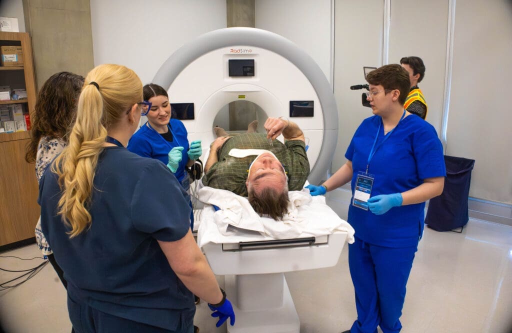 A second-year MRI student watches as two first-years wearing scrubs and gloves help an injured patient through an MRI machine.