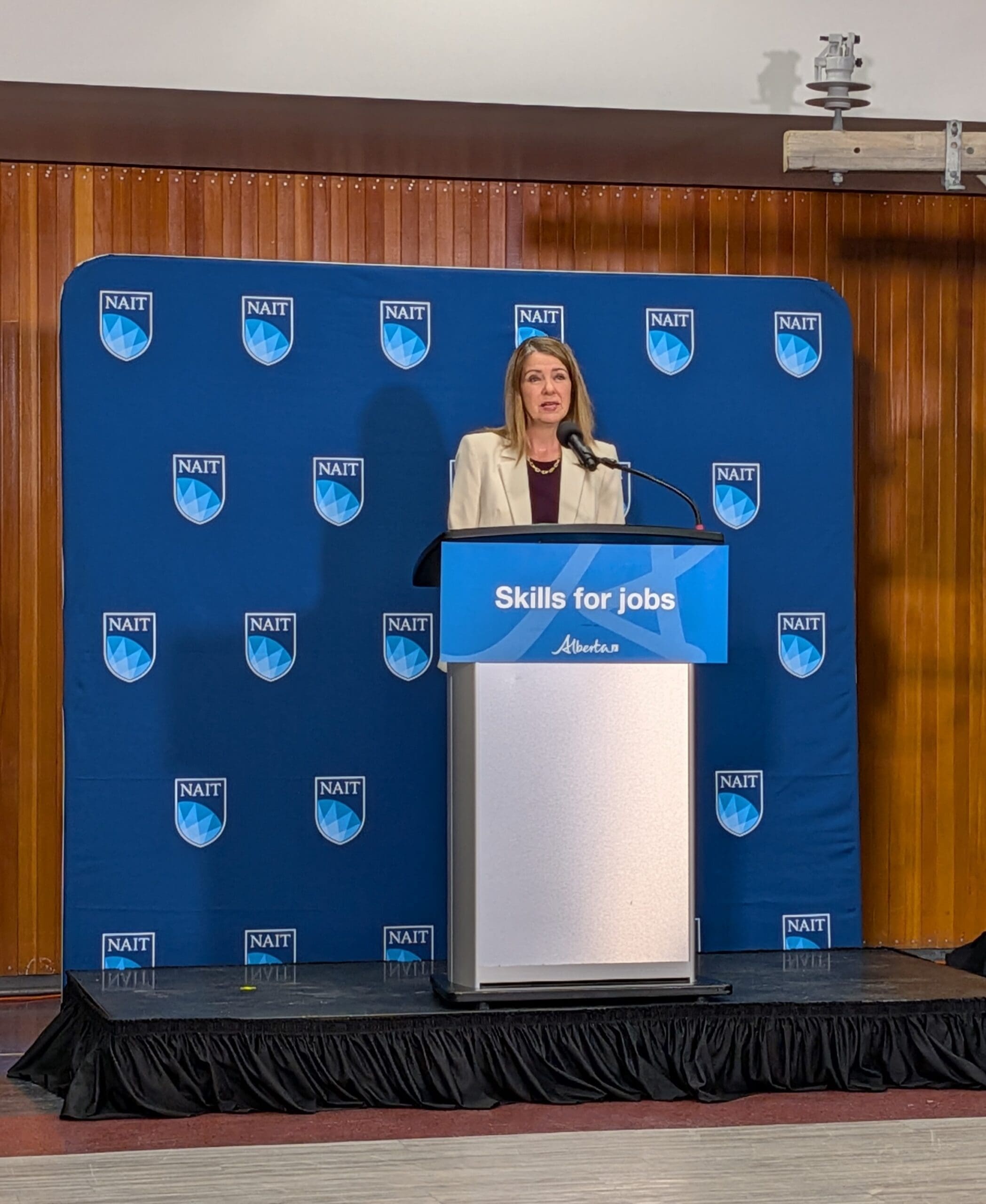 Premier Danielle Smith speaks into a microphone on a podium that reads "Skills for Jobs" with the Alberta government logo. The background is blue with NAIT's logo.